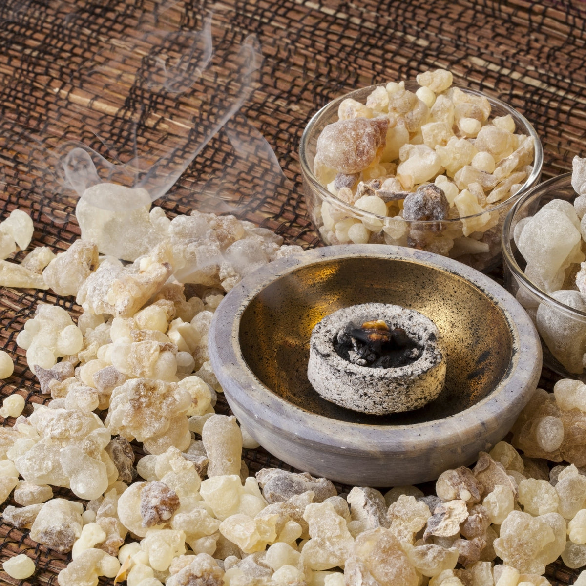 Frankincense resin pieces surround a small incense burner with a piece of resin smouldering in the centre, showing frankincense being used as incense.