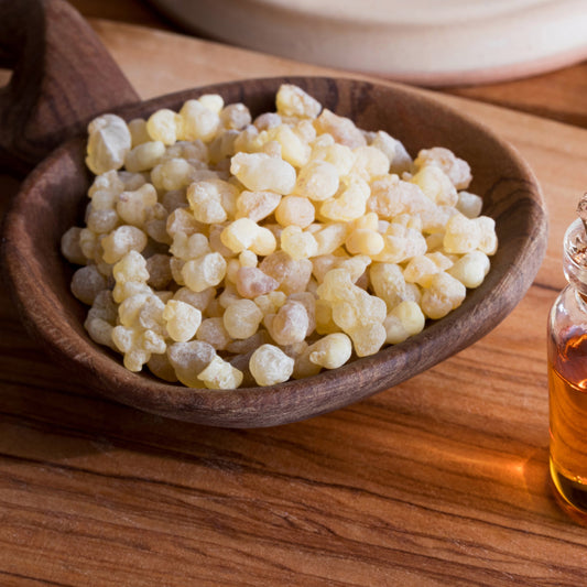 Pale golden frankincense resin pieces fill a wooden spoon on a timber surface, showing the irregular shape and natural translucent colouring of the resin.