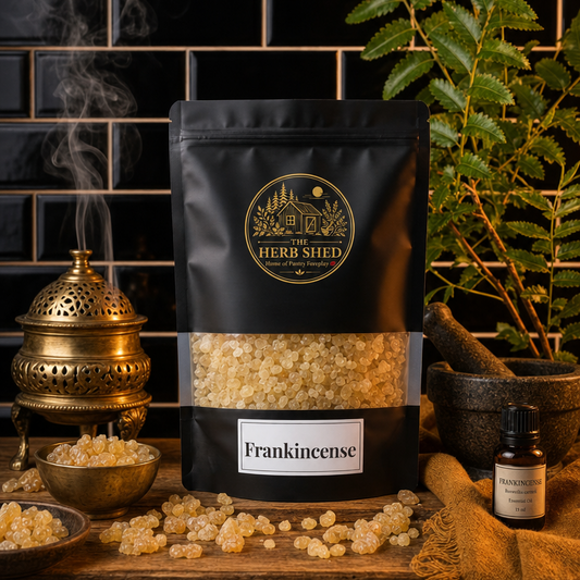 A black stand-up pouch of The Herb Shed Frankincense sits on a wooden surface with loose pale frankincense resin pieces, a brass incense burner, small bowl and essential oil bottle, surrounded by green foliage against a black tiled background.