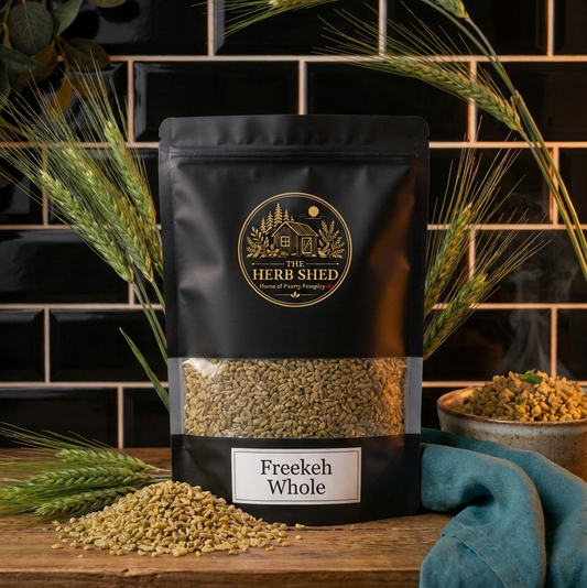 A black stand-up pouch of The Herb Shed Freekeh Whole in Wollongong, Australia, sits on a wooden surface with loose freekeh and wheat stems, surrounded by greenery and teal fabric against a black tiled background.