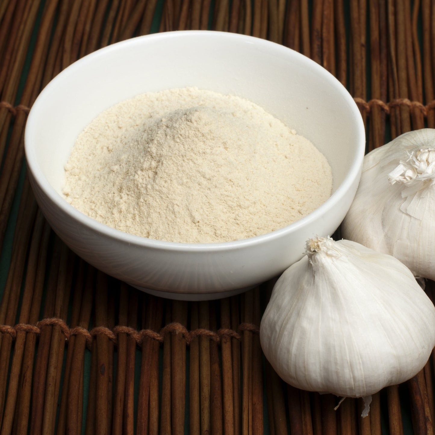 Fine garlic powder fills a white bowl beside a whole garlic bulb, showing the smooth, finely ground texture and pale cream colour of the powder.