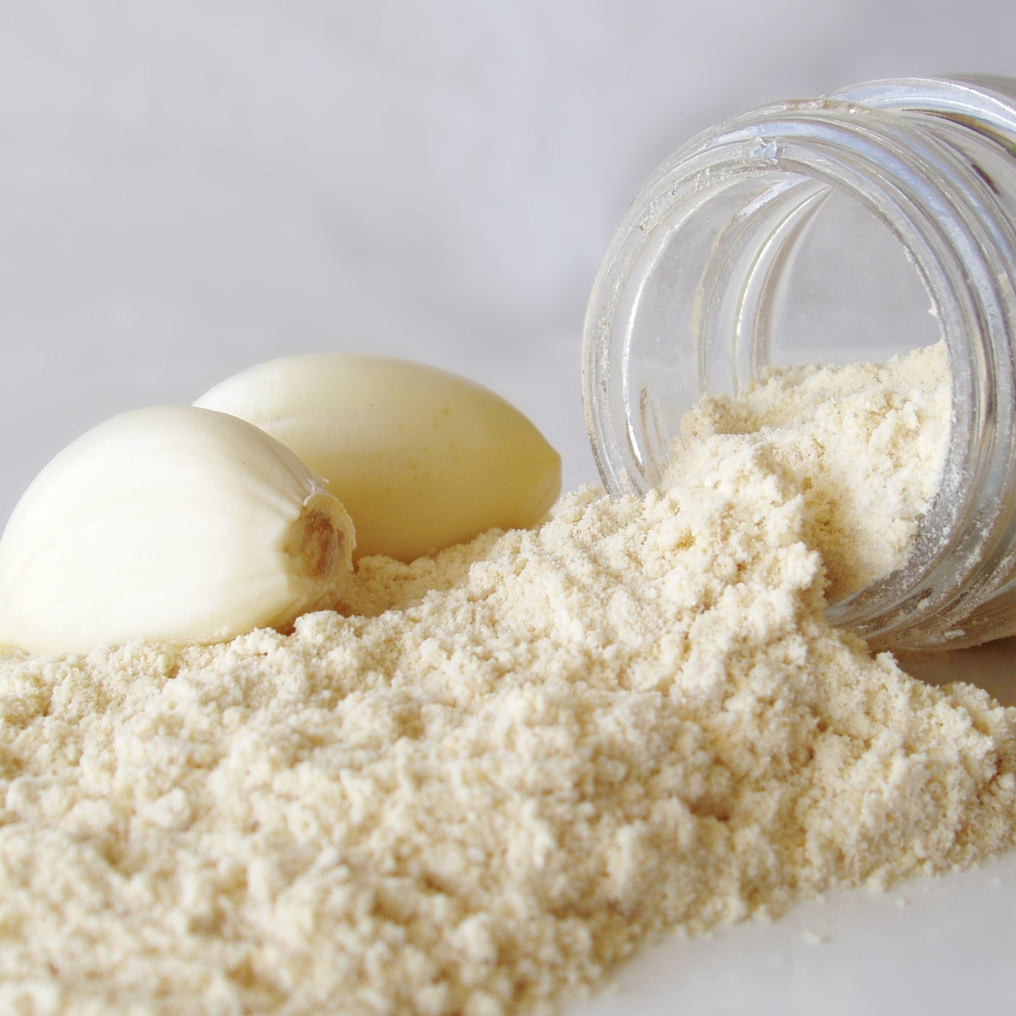 Fine garlic powder is piled beside fresh garlic cloves and a small glass jar, showing the pale cream colour and finely ground texture of the powder.