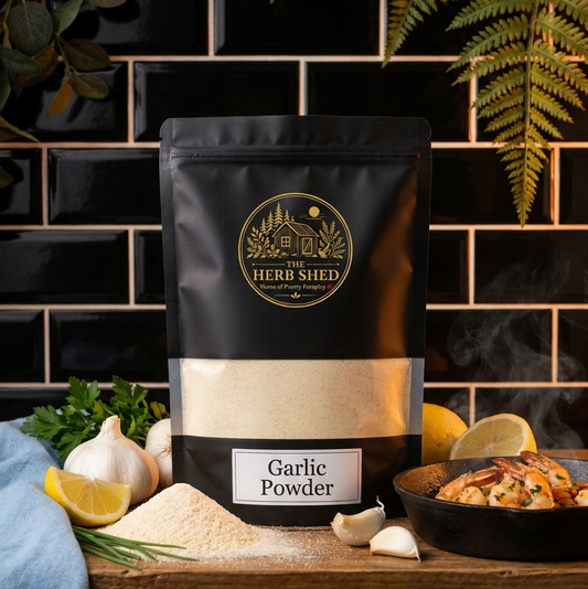 A black stand-up pouch of The Herb Shed Garlic Powder sits on a wooden kitchen surface with loose garlic powder, whole garlic bulbs, garlic cloves, lemon and fresh herbs, with a cooked dish beside it against a black tiled background.