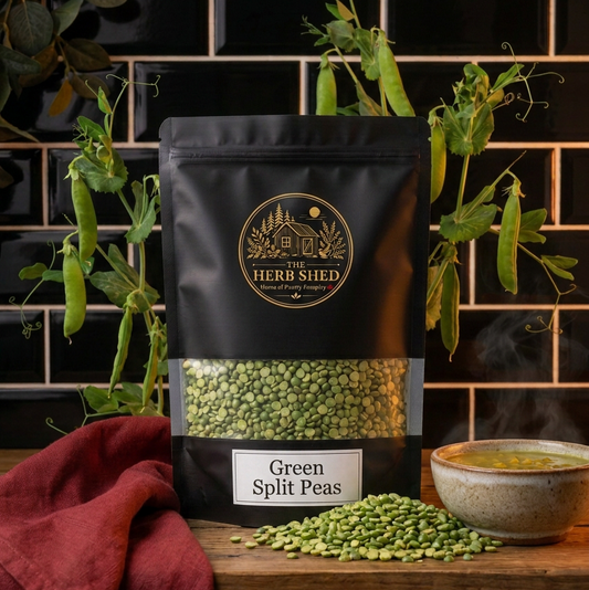 A black stand-up pouch of The Herb Shed Green Split Peas in Wollongong, Australia, sits on a wooden surface with loose split peas and a bowl of soup, surrounded by pea plants and deep red fabric against a black tiled background.