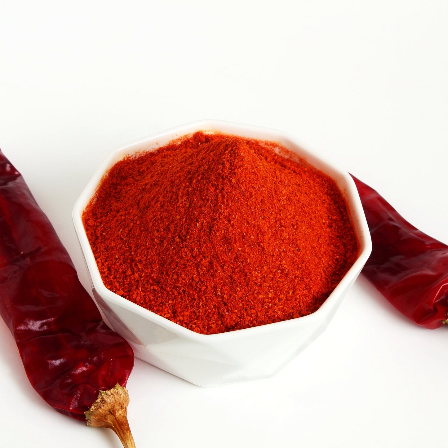 Bright red Kashmiri chilli powder fills a small white bowl with whole dried red chillies arranged beside it, showing the rich colour and fine texture of the ground spice.