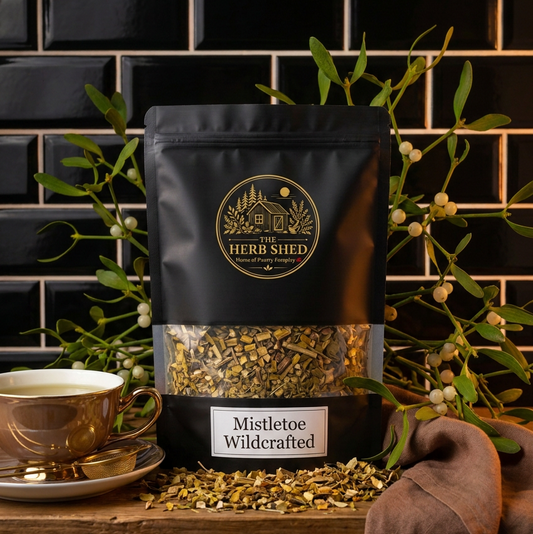 A black stand-up pouch of The Herb Shed Mistletoe Wildcrafted in Wollongong, Australia, sits on a wooden surface with loose dried mistletoe and a cup of herbal tea, surrounded by mistletoe branches against a black tiled background.