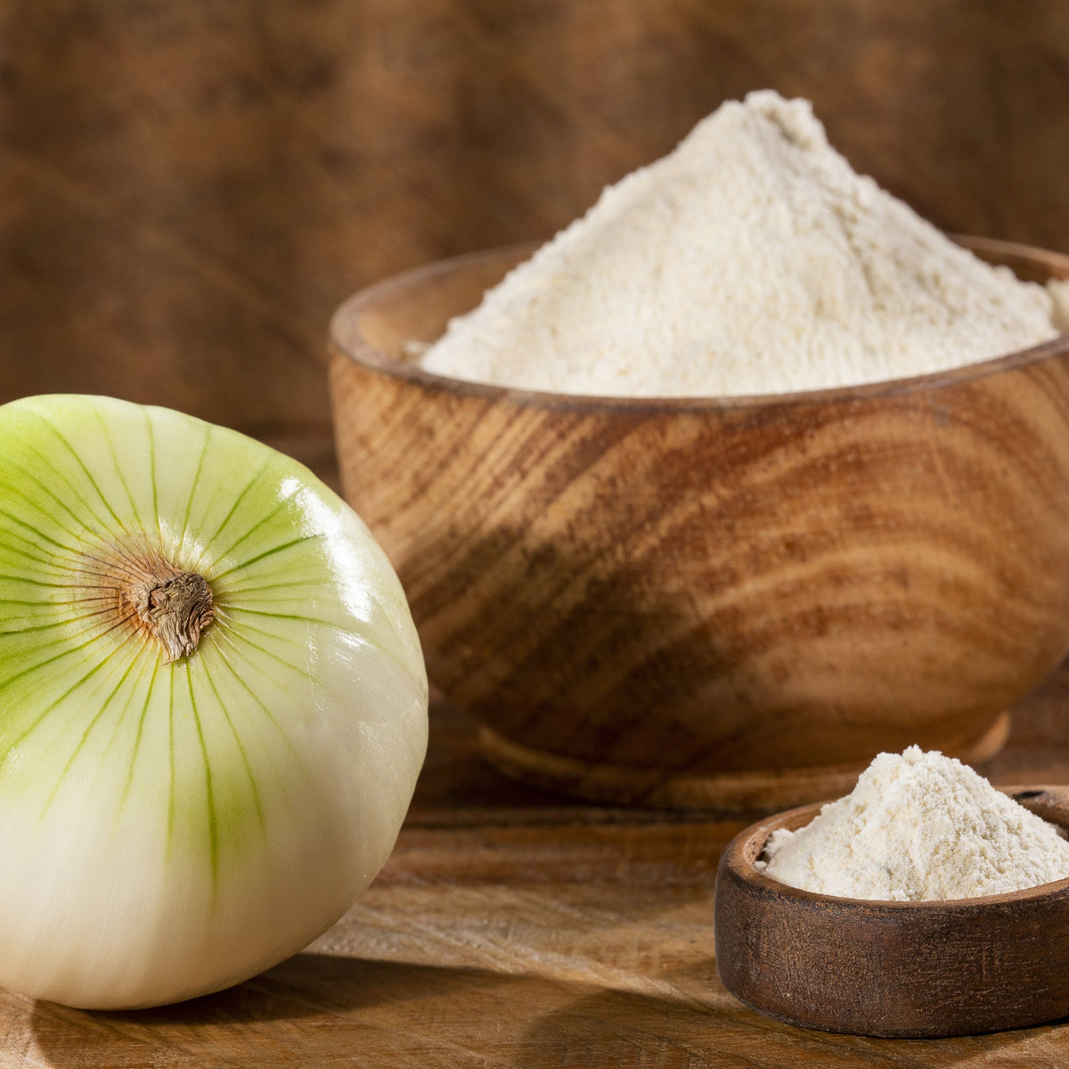 Fine onion powder is piled on a wooden board beside a whole fresh onion, with additional onion powder in a small wooden bowl.