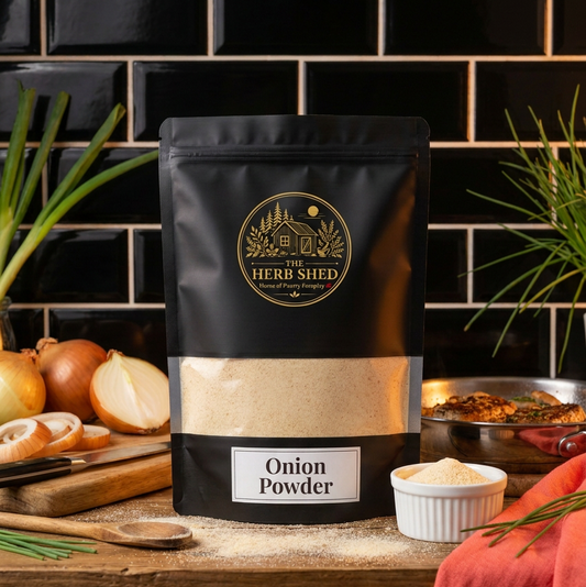 A black stand-up pouch of The Herb Shed Onion Powder sits on a wooden kitchen surface with loose onion powder, whole and cut brown onions, green onion leaves, a small white bowl and cooking ingredients, against a black tiled background with coral fabric.