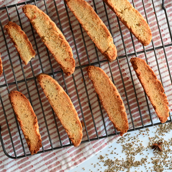Freshly baked biscotti are arranged on a cooling rack with loose aniseed scattered nearby, showing Organic Aniseed used in baking.