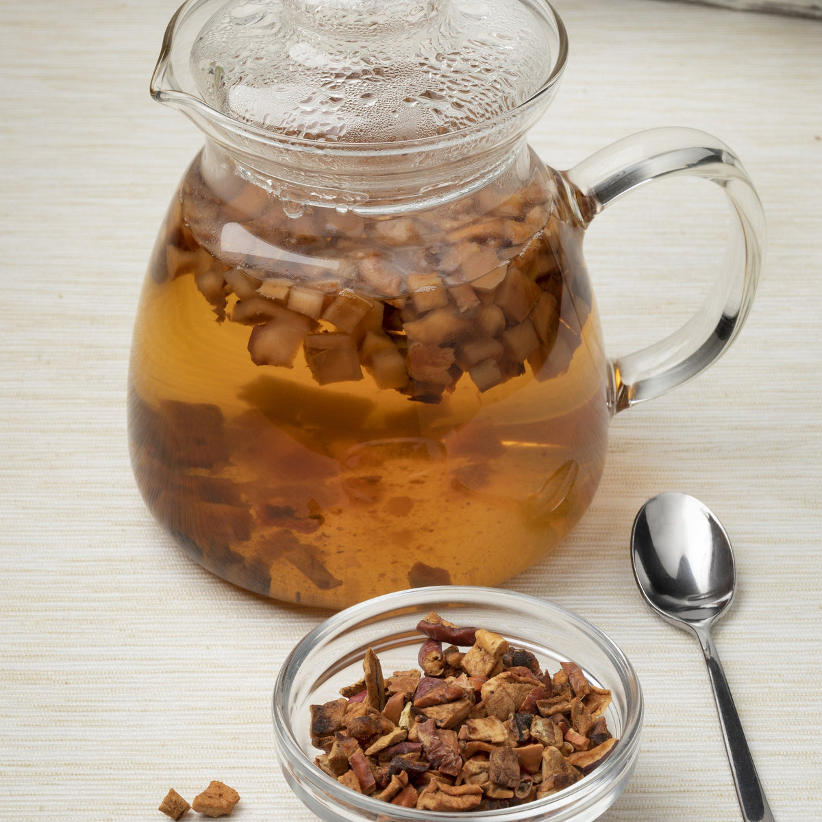 A glass pot of golden herbal tea brewed with dried apple pieces sits beside a small bowl of Organic Apple Pieces Dried and a teaspoon.