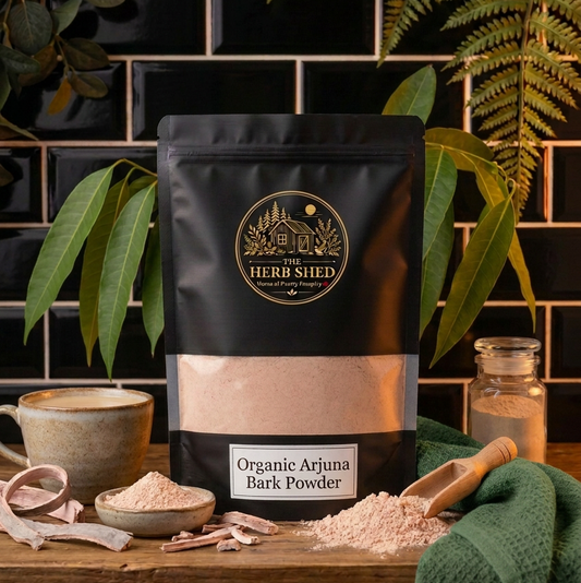A black stand-up pouch of The Herb Shed Organic Arjuna Bark Powder sits on a wooden surface with loose bark powder, pieces of dried bark, a wooden scoop and a cup, surrounded by green leaves and fabric against a black tiled background.