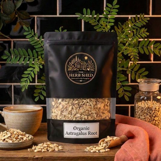 A black stand-up pouch of The Herb Shed Organic Astragalus Root sits on a wooden surface with dried astragalus root pieces, a small bowl and glass jar, surrounded by green foliage and soft rust-coloured fabric against a black tiled background.