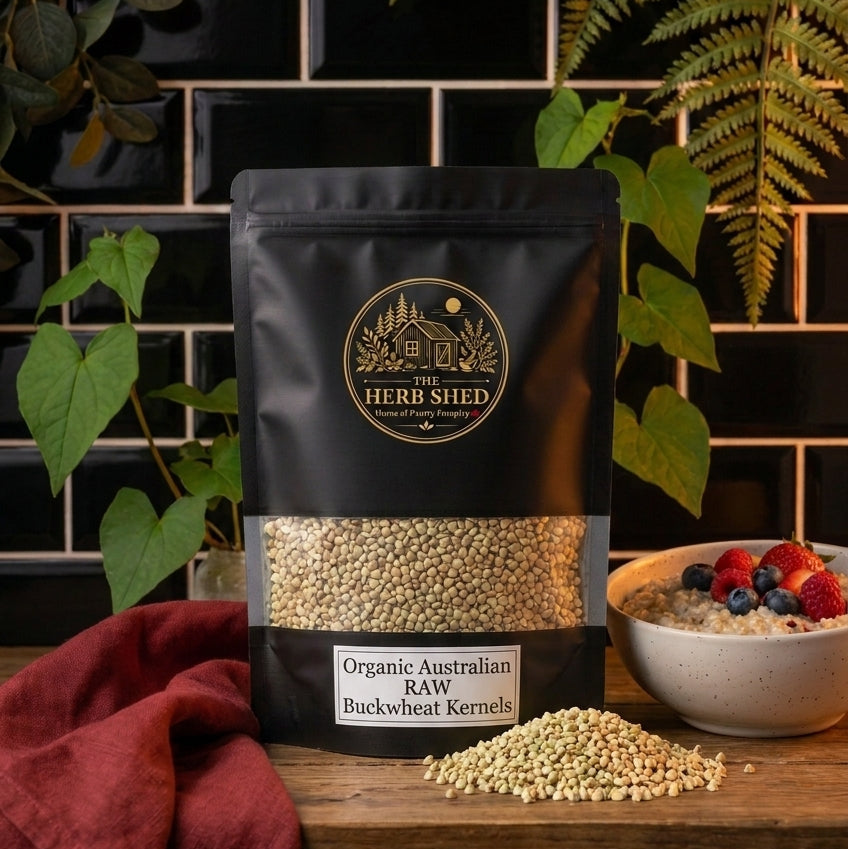 A black stand-up pouch of Organic Australian Raw Buckwheat Kernels displayed on a rustic wooden surface with scattered buckwheat kernels, green foliage, a bowl of berries and burgundy fabric against dark kitchen tiles, from The Herb Shed Wollongong Australia.