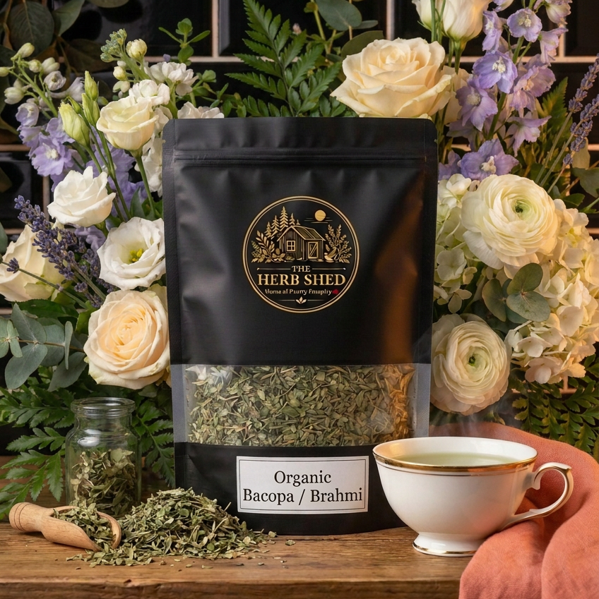 A black stand-up pouch of The Herb Shed Organic Bacopa / Brahmi sits on a wooden surface with loose dried herb, a small glass jar and a cup of herbal tea, surrounded by cream and purple flowers and leafy greenery against a black tiled background.