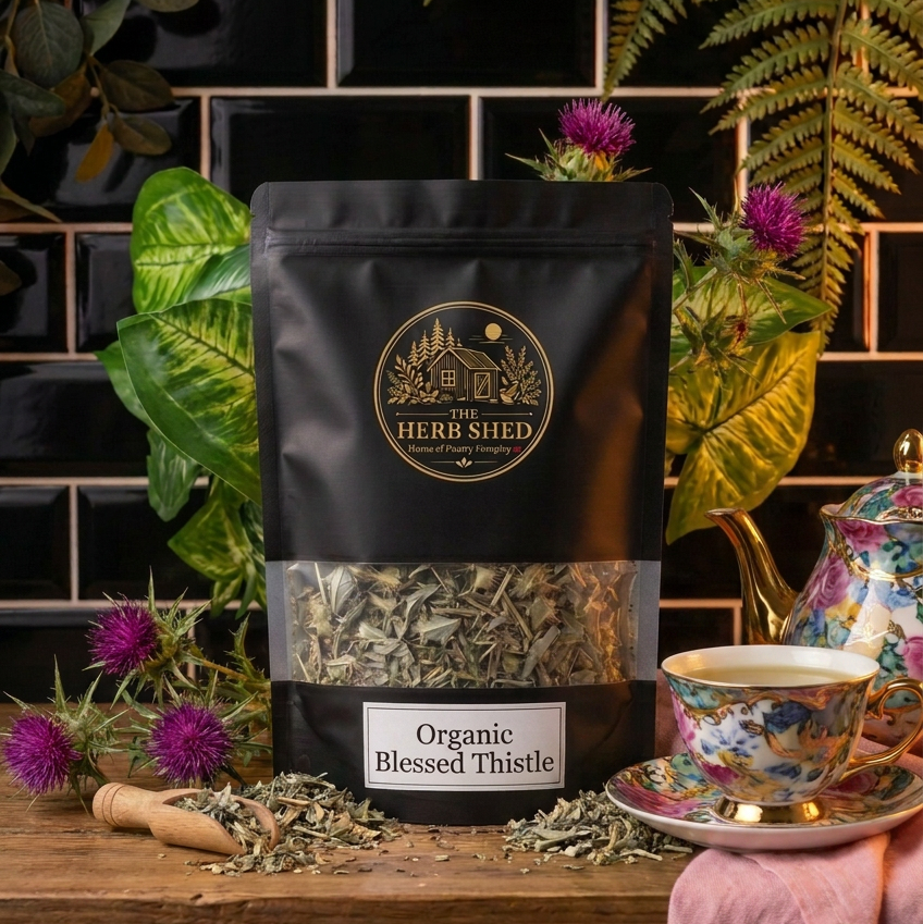 A black stand-up pouch of The Herb Shed Organic Blessed Thistle sits on a wooden surface with loose dried blessed thistle, a wooden scoop, purple thistle flowers and green foliage, with a decorative teacup against a black tiled background.