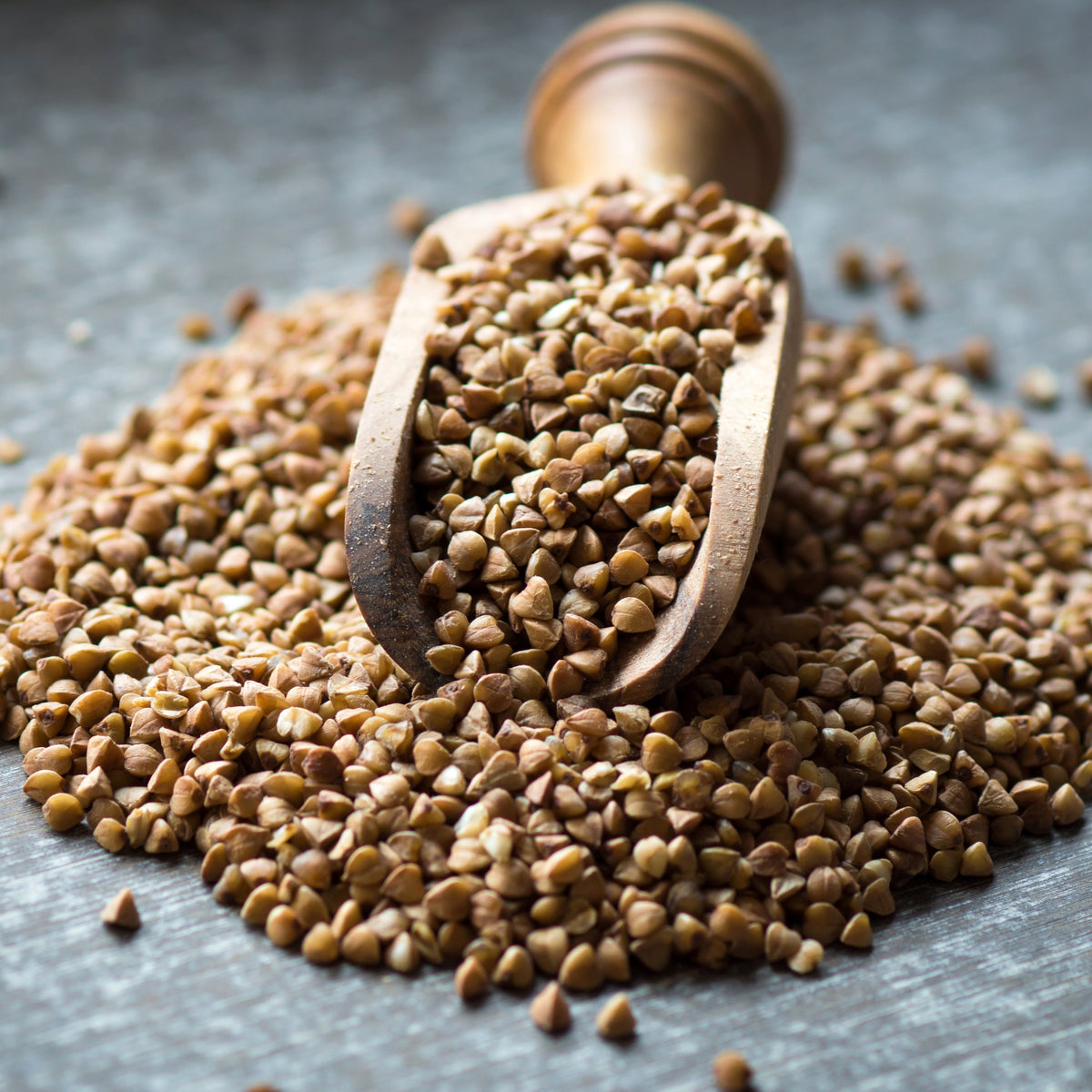 Organic buckwheat grains are piled on a textured surface with a small wooden scoop resting among them, showing their triangular shape and natural tan and brown colouring.