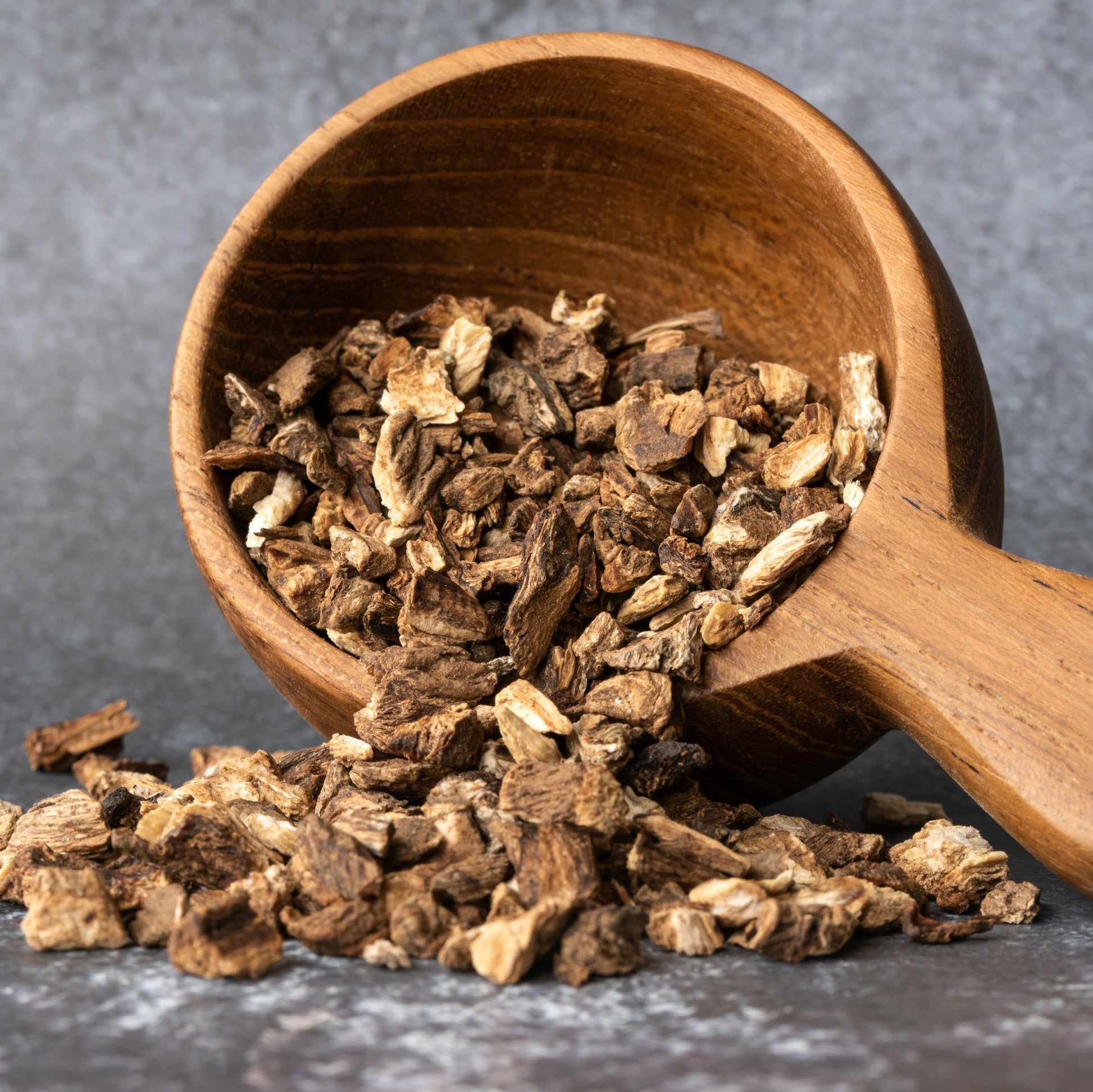 Dried organic burdock root pieces spill from a small wooden scoop onto a dark surface, showing their chopped shape and natural tan and brown colouring.