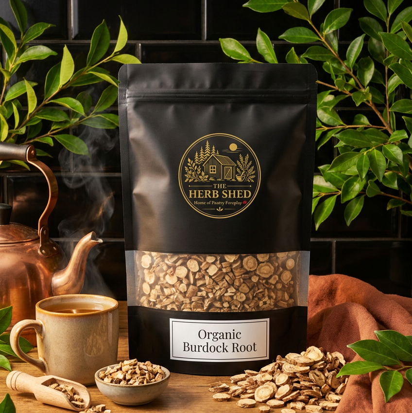 A black stand-up pouch of The Herb Shed Organic Burdock Root sits on a wooden surface with loose dried burdock root pieces, a small bowl, teacup, green foliage and copper kitchenware, against a black tiled background with warm brown fabric.