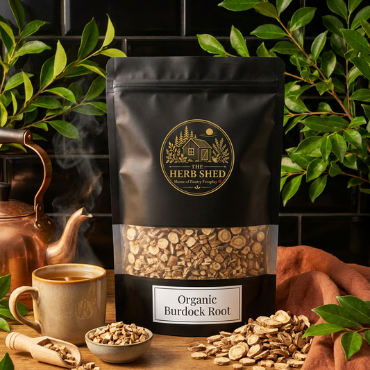 A black stand-up pouch of The Herb Shed Organic Burdock Root sits on a wooden surface with loose dried burdock root pieces, a small bowl, teacup, green foliage and copper kitchenware, against a black tiled background with warm brown fabric.