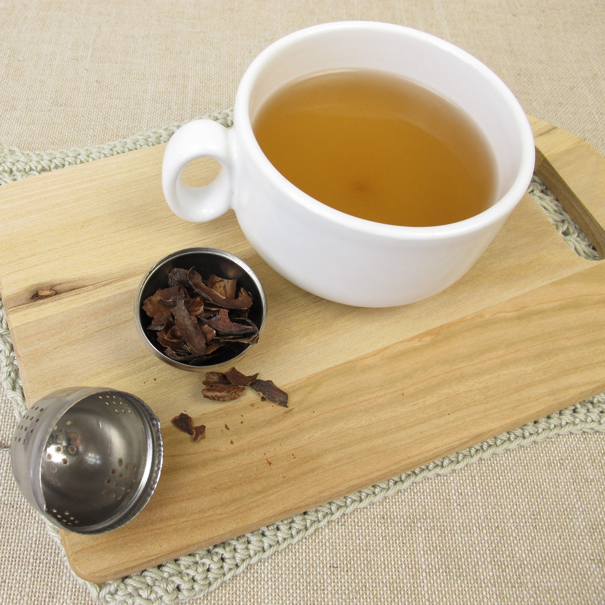 A white cup of golden brewed Cacao Husk Tea sits on a wooden board beside loose cacao husks in a small tea infuser and a metal teaspoon.