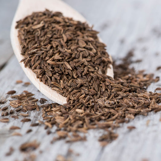 Organic caraway seeds are scattered across a light surface, showing their elongated curved shape, fine ridges and natural brown colouring.