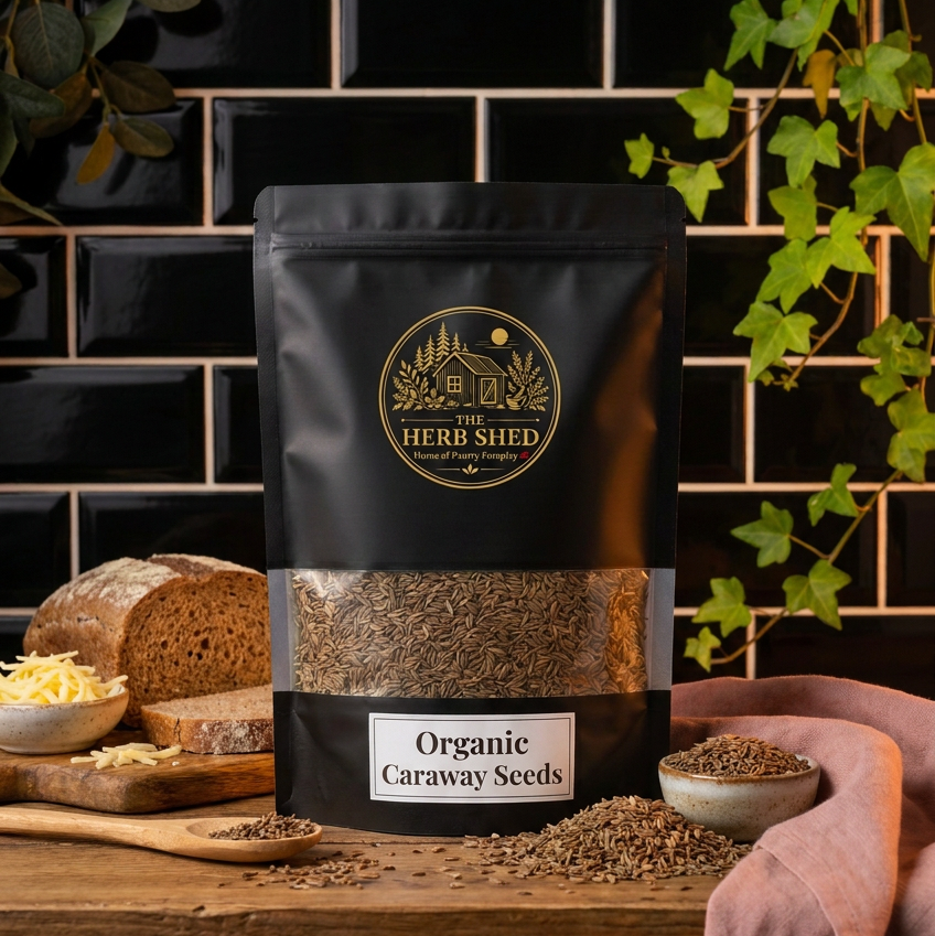 A black stand-up pouch of The Herb Shed Organic Caraway Seeds sits on a wooden surface with loose caraway seeds, a small bowl of seeds, rustic bread and lemon, surrounded by green ivy against a black tiled background with soft pink fabric.