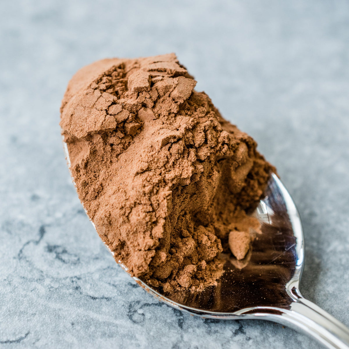 Organic roasted carob powder is heaped onto a metal spoon, showing its fine texture and rich warm brown colour.