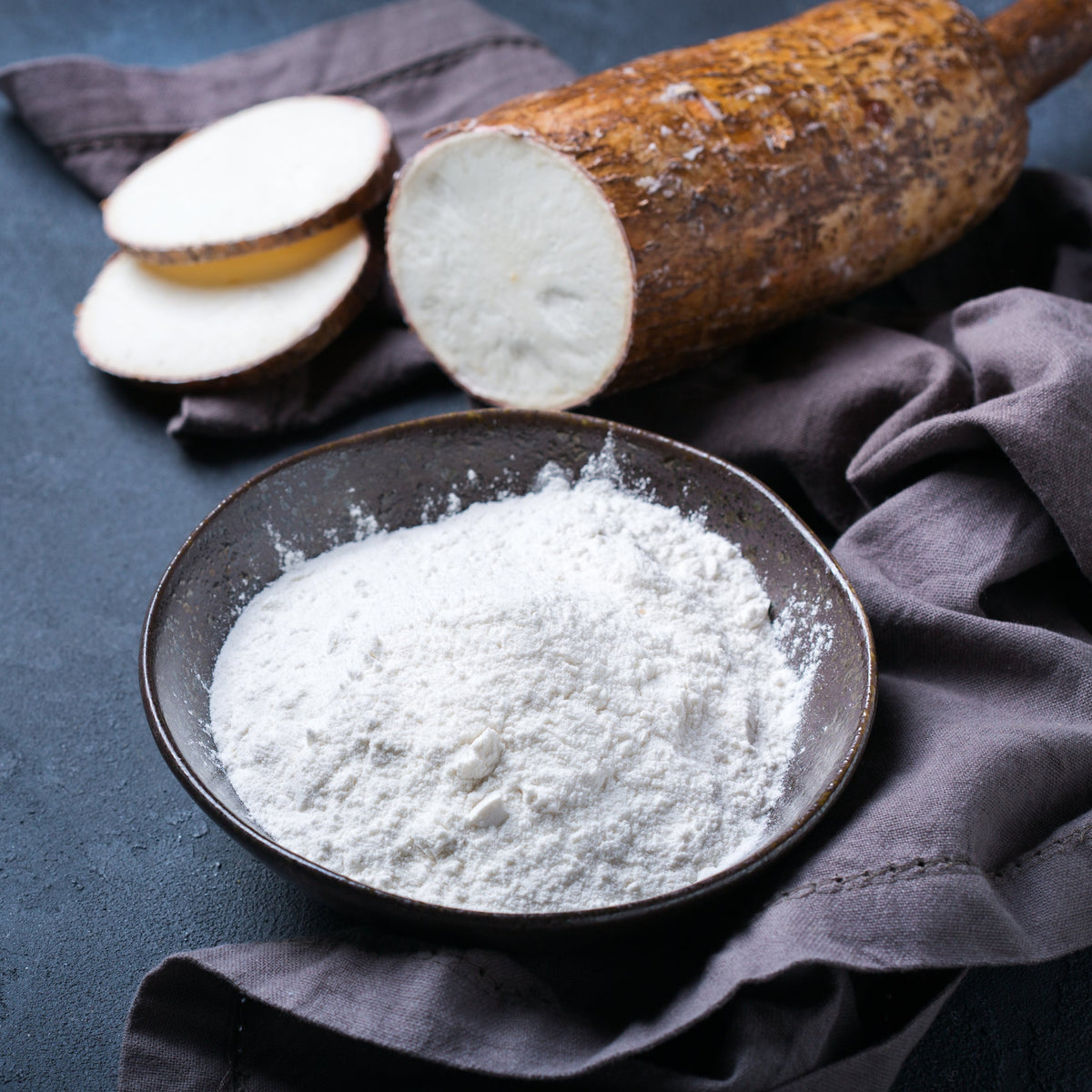 Organic Cassava Flour Extra Fine fills a dark bowl beside whole and cut cassava root, showing the finely milled flour alongside the root from which it is produced.