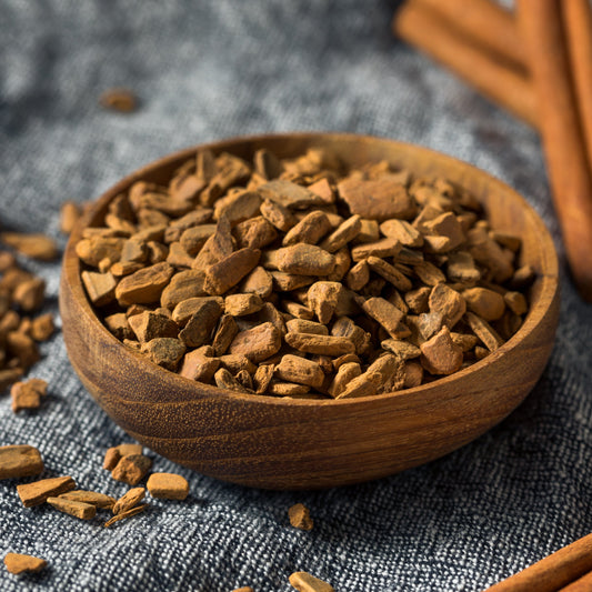 Organic Cassia Cinnamon Pieces fill a wooden bowl on a dark rustic surface, showing the chopped pieces of cinnamon bark and their natural warm brown colouring and woody texture.