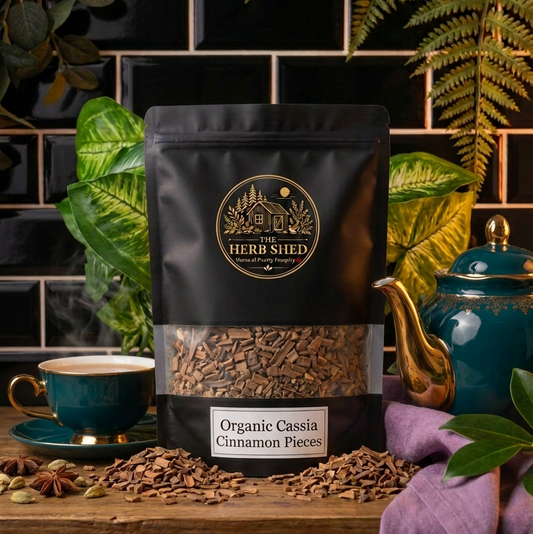 A black stand-up pouch of The Herb Shed Organic Cassia Cinnamon Pieces sits on a wooden surface with loose cinnamon pieces, whole spices and a cup of tea, surrounded by leafy greenery, a teal teapot and soft purple fabric against a black tiled background.