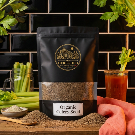 A black stand-up pouch of The Herb Shed Organic Celery Seed sits on a wooden surface with loose celery seeds, fresh celery stalks and leaves, a small bowl of seeds and a glass of tomato juice, against a black tiled background with soft coral fabric.