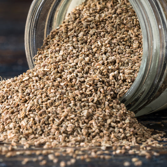 Organic celery seeds spill from a glass jar onto a dark surface, showing their tiny oval shape, fine texture and natural brown colouring.