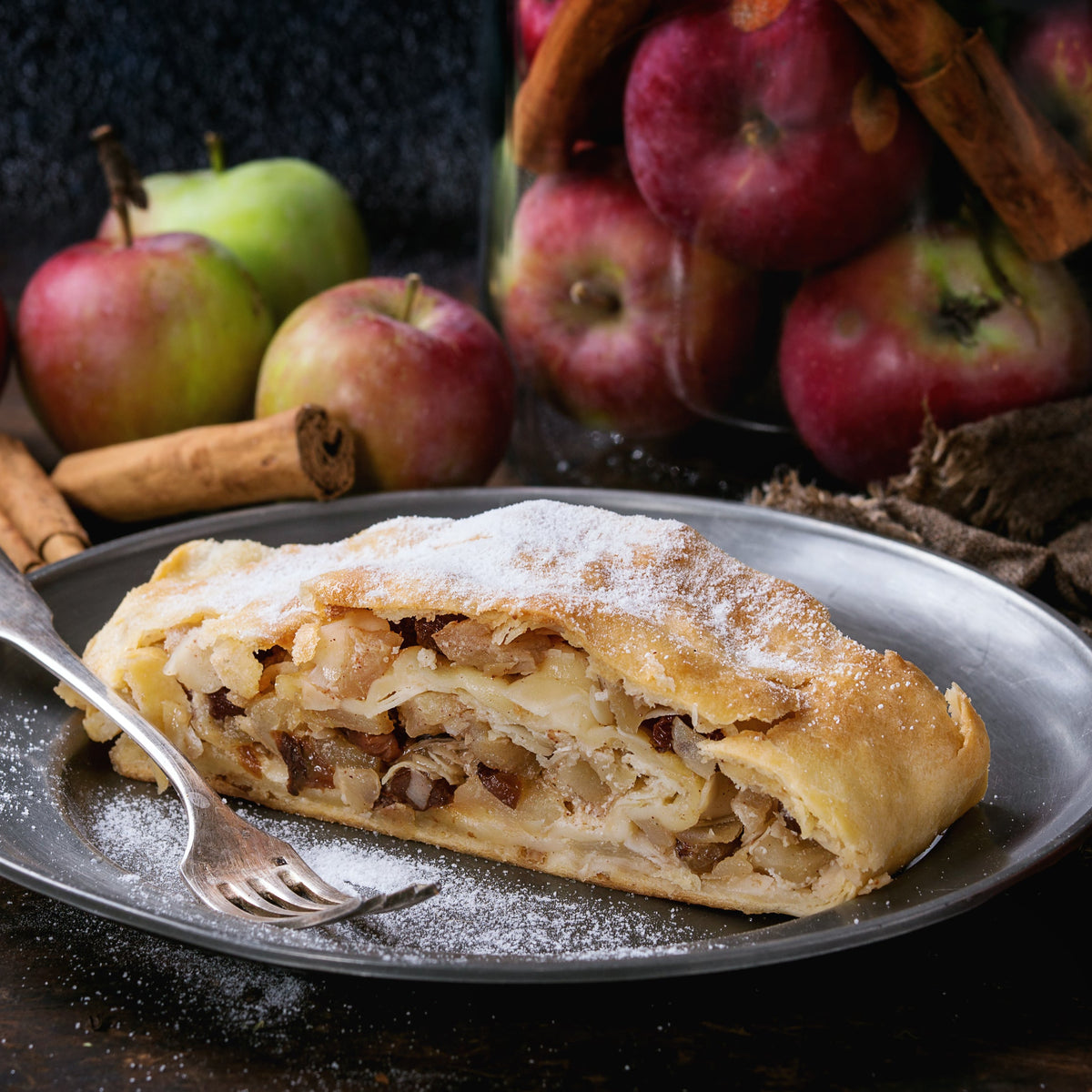 A slice of apple strudel filled with spiced apple sits beside fresh apples, showing Organic Ceylon Cinnamon Powder used in sweet baking.