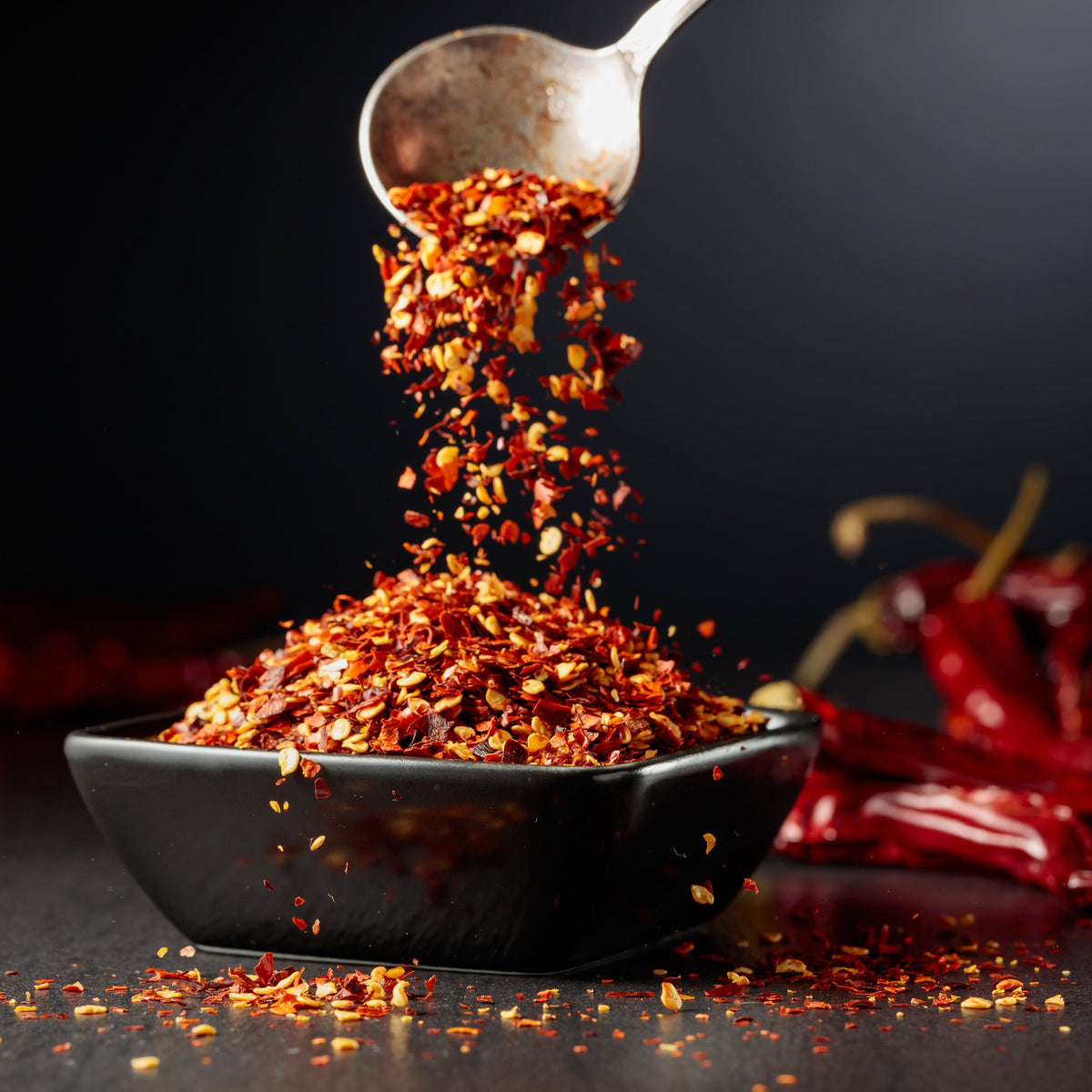 Organic Chilli Flakes are sprinkled from a spoon into a small cooking pot, with dried red chillies and loose flakes surrounding the dish.