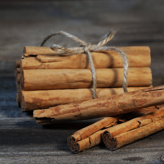 Whole Organic Cinnamon Sticks are bundled with twine on a dark rustic surface, showing the rolled bark and its natural warm brown colouring and woody texture.