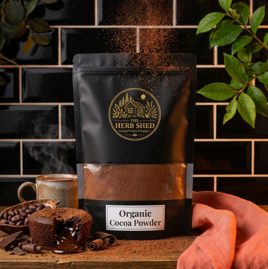 A black stand-up pouch of The Herb Shed Organic Cocoa Powder sits on a wooden surface with loose cocoa powder, cocoa beans, chocolate cake and a ceramic mug, surrounded by green foliage and warm terracotta fabric against a black tiled background.