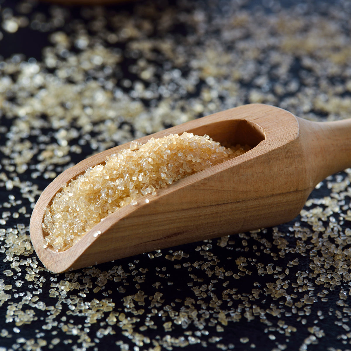 Organic coconut sugar fills a wooden scoop with loose granules scattered around it, showing the fine texture and natural golden-brown colour of the sugar.