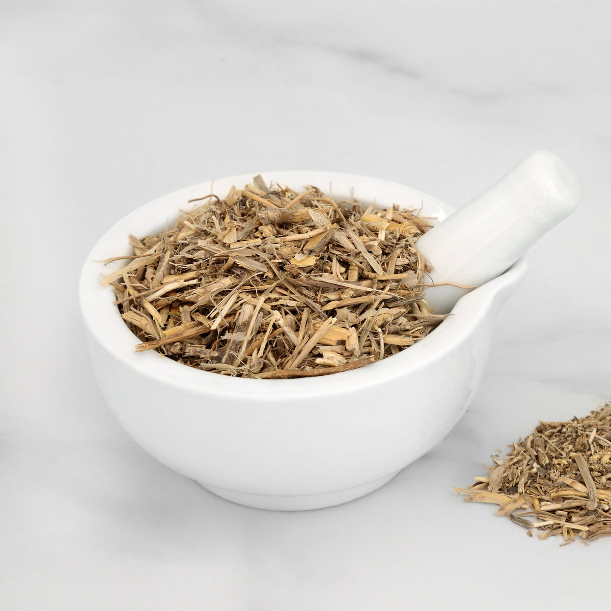 Organic dried couch grass root fills a small white bowl with loose pieces scattered beside it, showing the thin cut texture and natural pale tan colouring of the dried root.