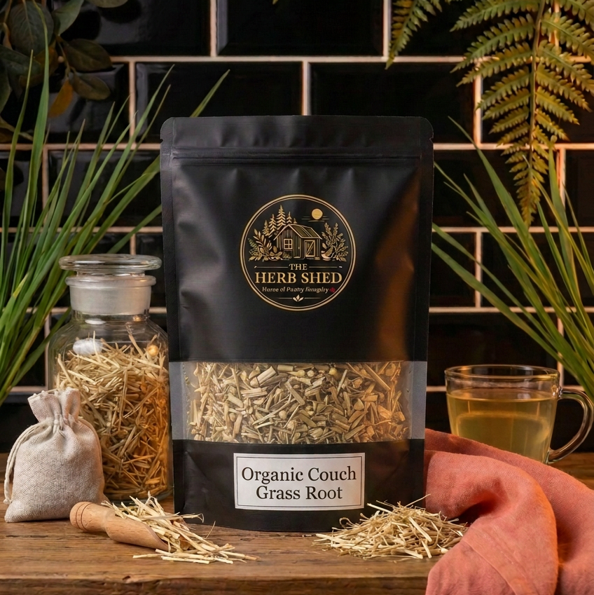 A black stand-up pouch of The Herb Shed Organic Couch Grass Root sits on a wooden surface with loose dried couch grass root, a glass jar of dried root, a small cloth bag and a cup of brewed tea, surrounded by green foliage against a black tiled background with soft terracotta fabric.