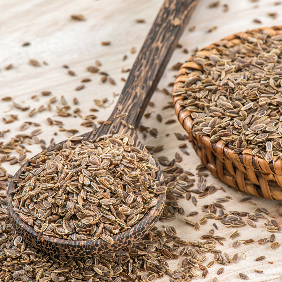 Organic dill seeds are scattered across a light surface beside a metal scoop filled with dill seeds, showing their small, flat and ridged texture.
