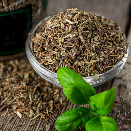 A close-up of dried mixed herbs in a small bowl, showing the blend of finely cut green and brown dried leaves, with fresh green herb leaves beside the bowl.