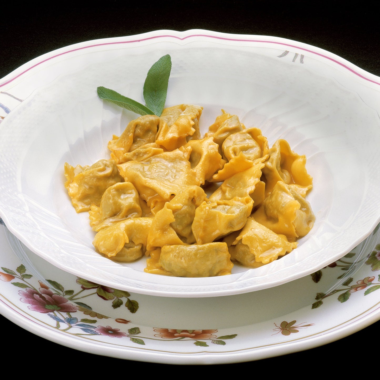 Pasta with pieces of meat and dried sage is served in a white ceramic bowl on a patterned plate.