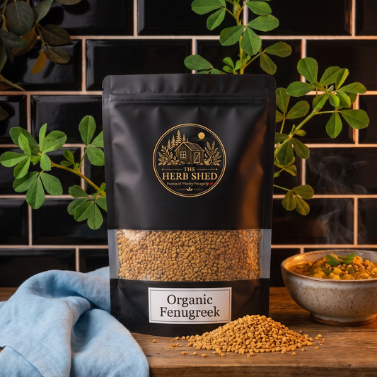 A black stand-up pouch of The Herb Shed Organic Fenugreek in Wollongong, Australia, sits on a wooden surface with loose fenugreek seeds and a bowl of cooked food, surrounded by green fenugreek leaves and soft blue fabric against a black tiled background.