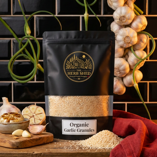 A black stand-up pouch of The Herb Shed Organic Garlic Granules sits on a wooden surface with loose garlic granules, whole garlic bulbs, garlic cloves and green garlic stems, against a black tiled background with deep red fabric.