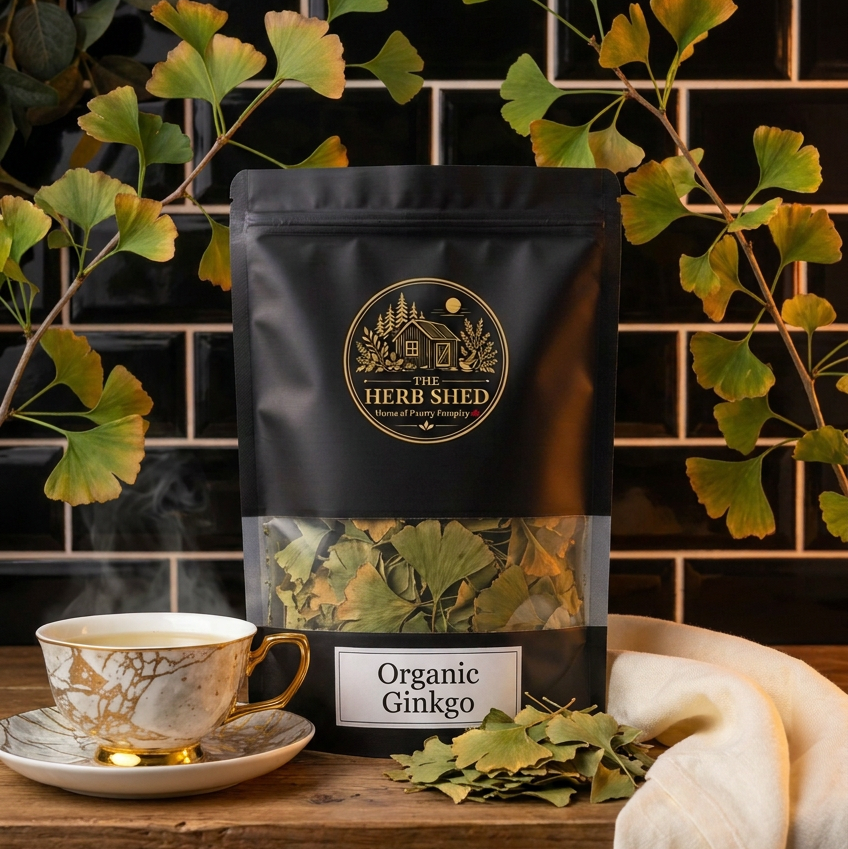 A black stand-up pouch of The Herb Shed Organic Ginkgo in Wollongong, Australia, sits on a wooden surface with loose dried ginkgo leaves and a teacup, surrounded by distinctive fan-shaped ginkgo leaves against a black tiled background.