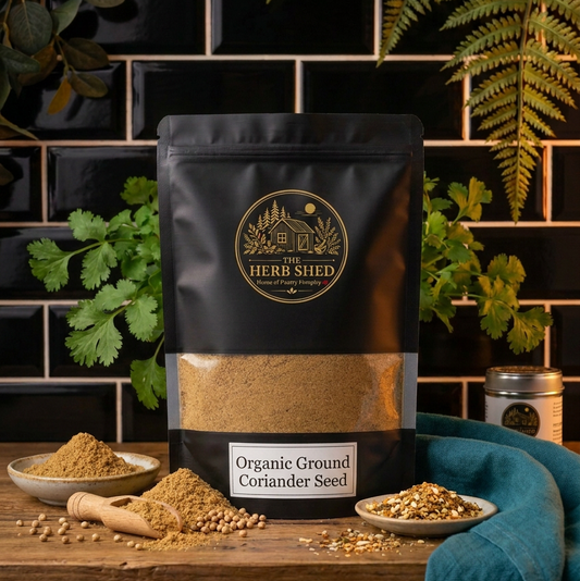 A black stand-up pouch of The Herb Shed Organic Ground Coriander Seed sits on a wooden surface with ground coriander, whole coriander seeds and a wooden scoop, surrounded by fresh coriander leaves and soft teal fabric against a black tiled background.