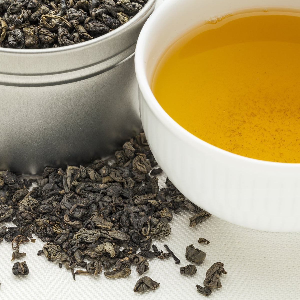 A cup of golden brewed Gunpowder Green Tea sits beside loose organic rolled green tea leaves and a container of tea, showing the dried leaves alongside the prepared infusion.