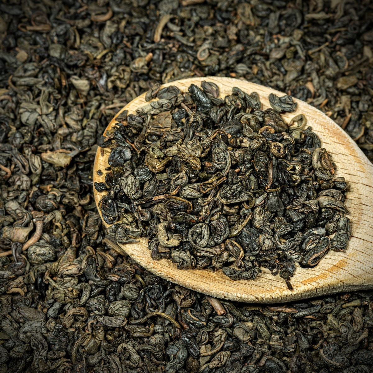 Organic Gunpowder Green Tea leaves fill a wooden scoop surrounded by loose tea, showing the small tightly rolled leaves and natural dark green colouring.