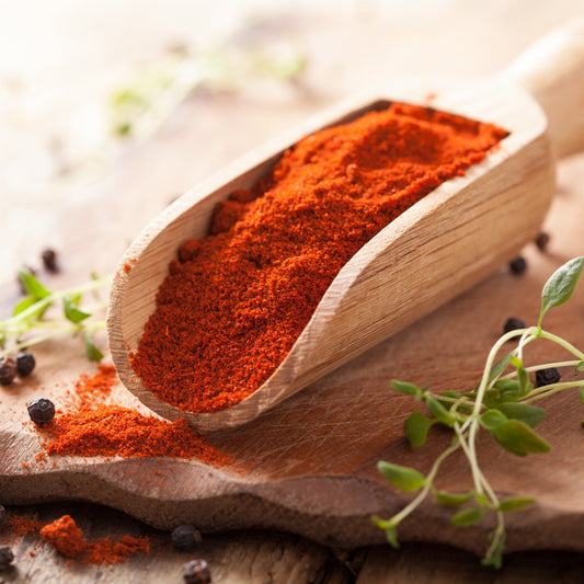 Hot paprika powder fills a wooden scoop on a rustic surface, showing the fine texture and rich red-orange colour of the ground spice.