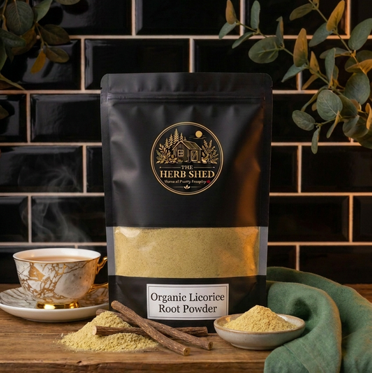 A black stand-up pouch of The Herb Shed Organic Licorice Root Powder in Wollongong, Australia, sits on a wooden surface with loose powder, licorice root pieces and a teacup, surrounded by greenery against a black tiled background.